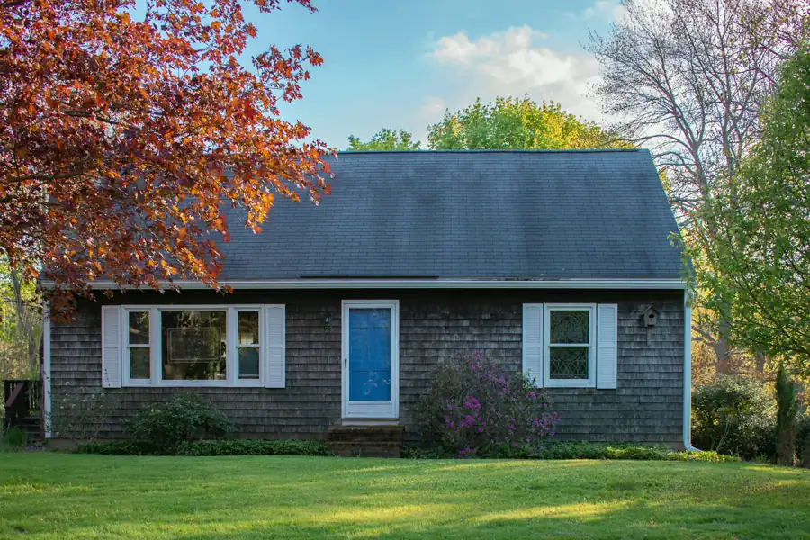 A grey cedar shingle Cape with a blue front door and white shutters sits on a green lawn framed by spring trees.