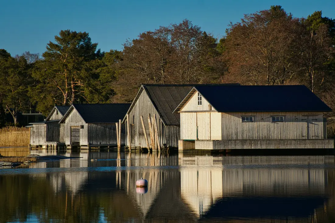 Weathered wooden boathouses stand at the edge of a still bay, their reflections doubled in the water below a line of trees.