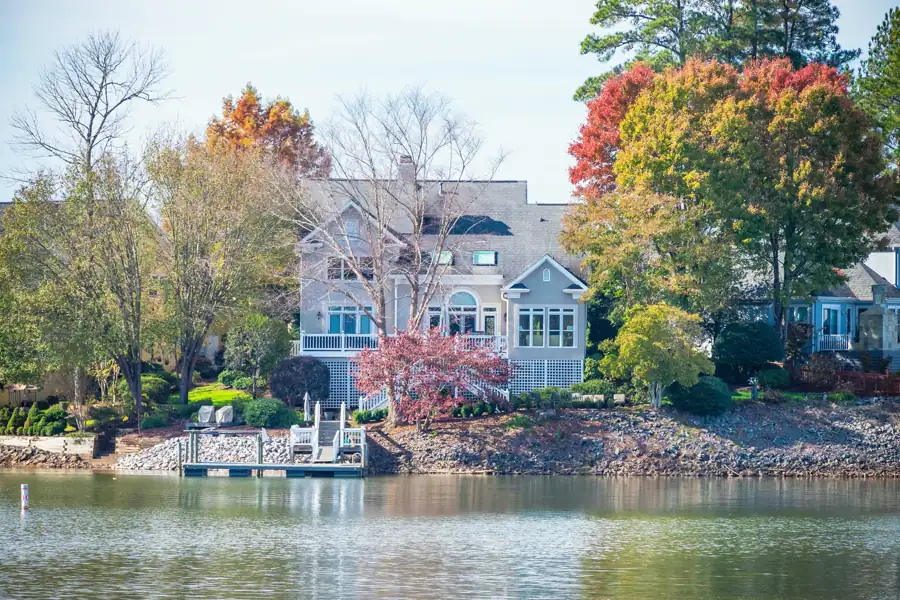 A pale grey lake house with a wide porch faces the water above a rock shoreline and a private dock, framed by autumn trees.