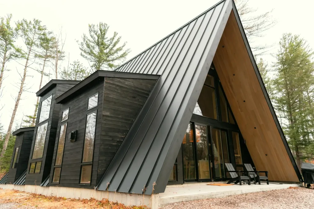 A black A-frame house with a standing seam metal roof, tall windows and two Adirondack chairs on the patio sits among pines.