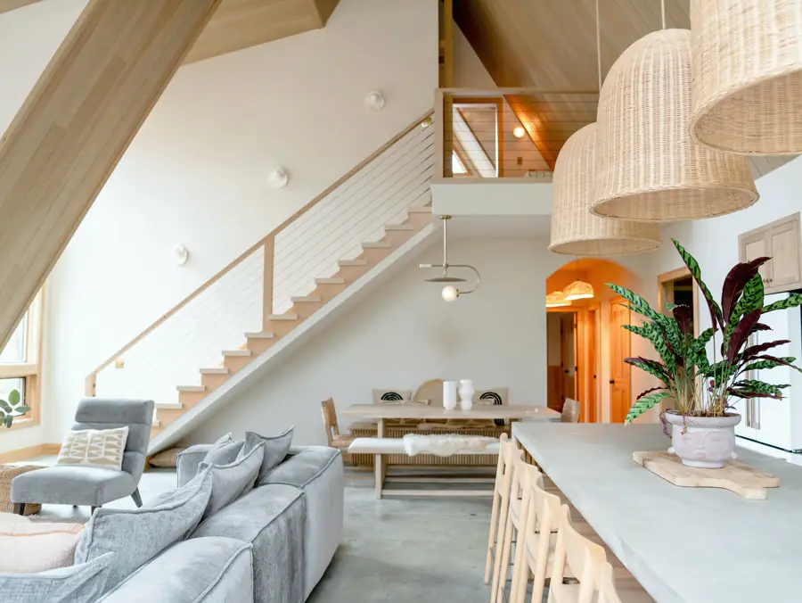 A long concrete dining table with oak chairs sits beside the living area, where a light wood stair climbs to an open loft.