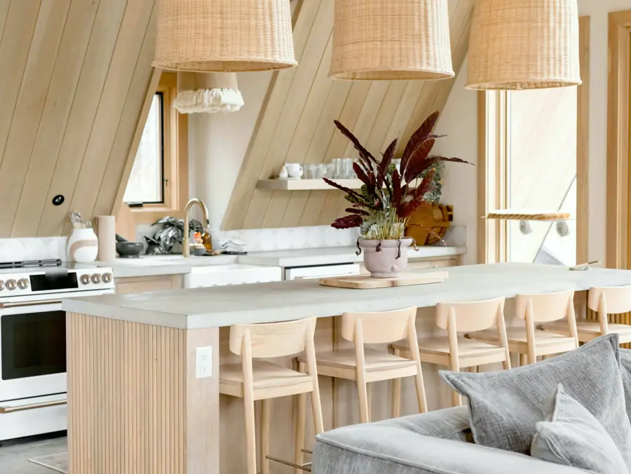 An open kitchen with a concrete island, pale oak stools, rattan pendant lights and a white range under a sloping wood ceiling.