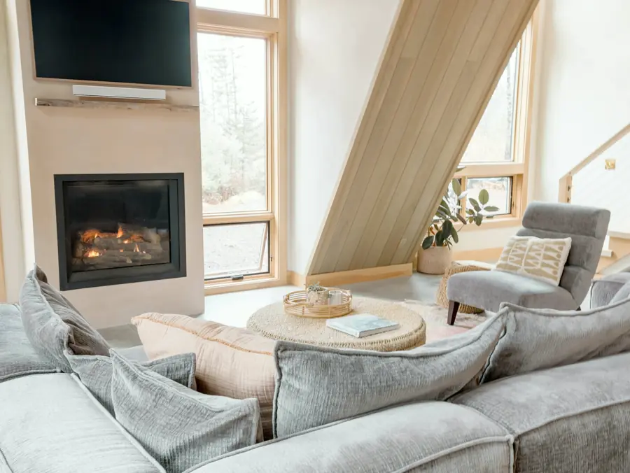 A bright living room under a pale wood A-frame ceiling, with a deep grey sectional, a woven coffee table and a gas fireplace.
