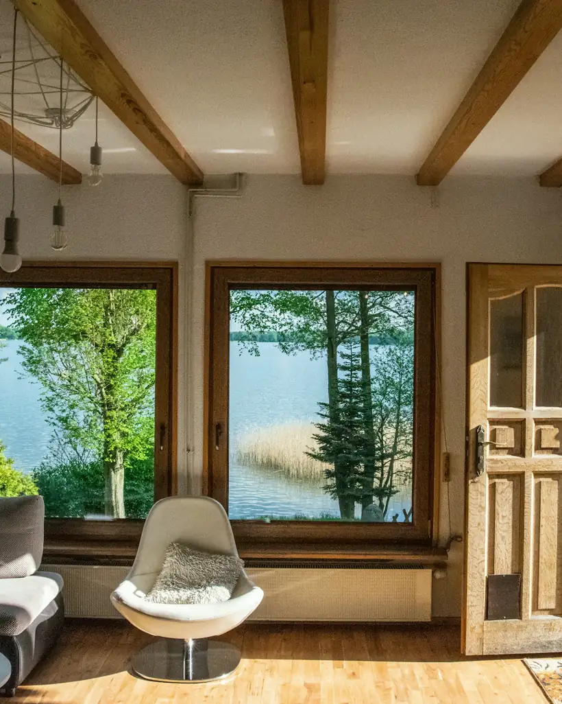 A sunlit living room with exposed wood beams, a grey sofa and a white swivel chair looks out through two windows to a lake.