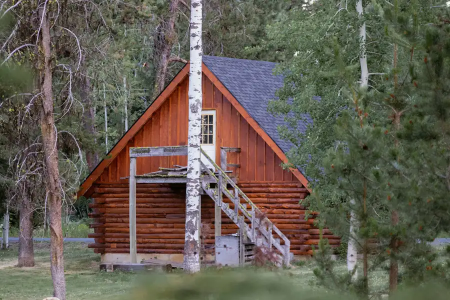 A log cabin with a red board and batten gable and an outside wooden stair stands among pines and aspens.