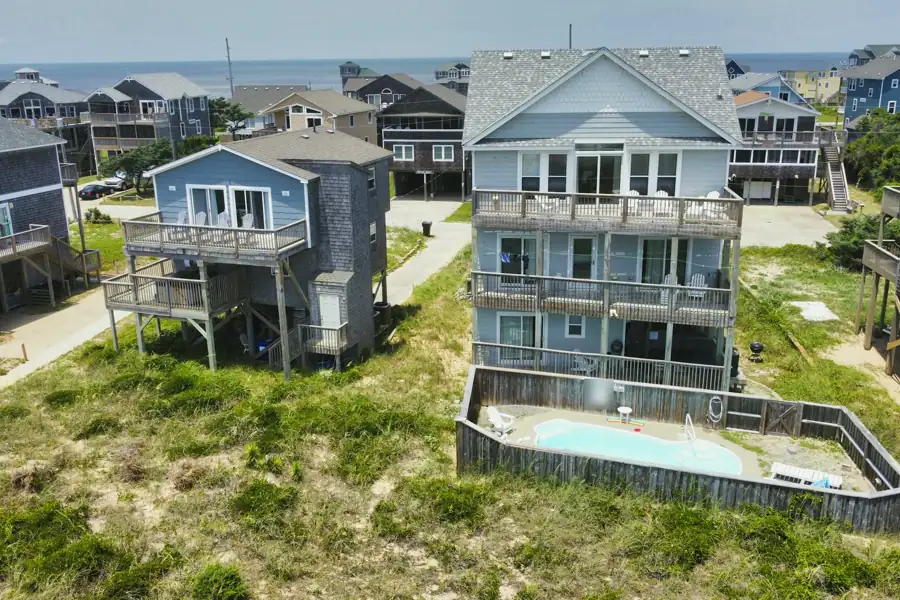 Raised beach houses with wide stacked decks stand on grassy dunes, the nearest with a fenced pool, and the ocean beyond.