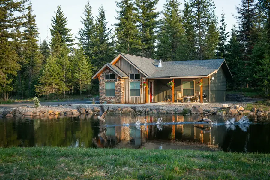 A small cottage in stone, metal and green siding sits beside the water among firs, with rocking chairs on its front porch.